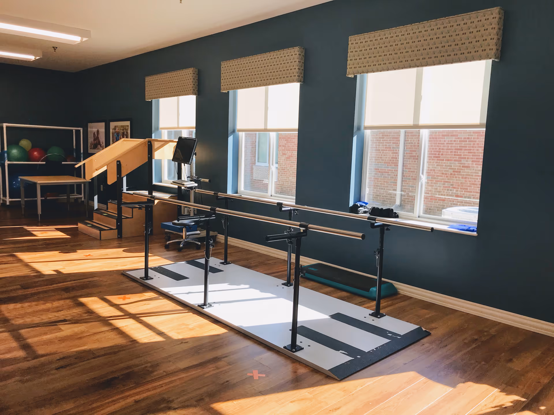 A rehabilitation room with parallel bars for walking exercises, a small set of wooden stairs with a handrail, exercise balls on a rack, a computer workstation, and large windows letting in natural light.