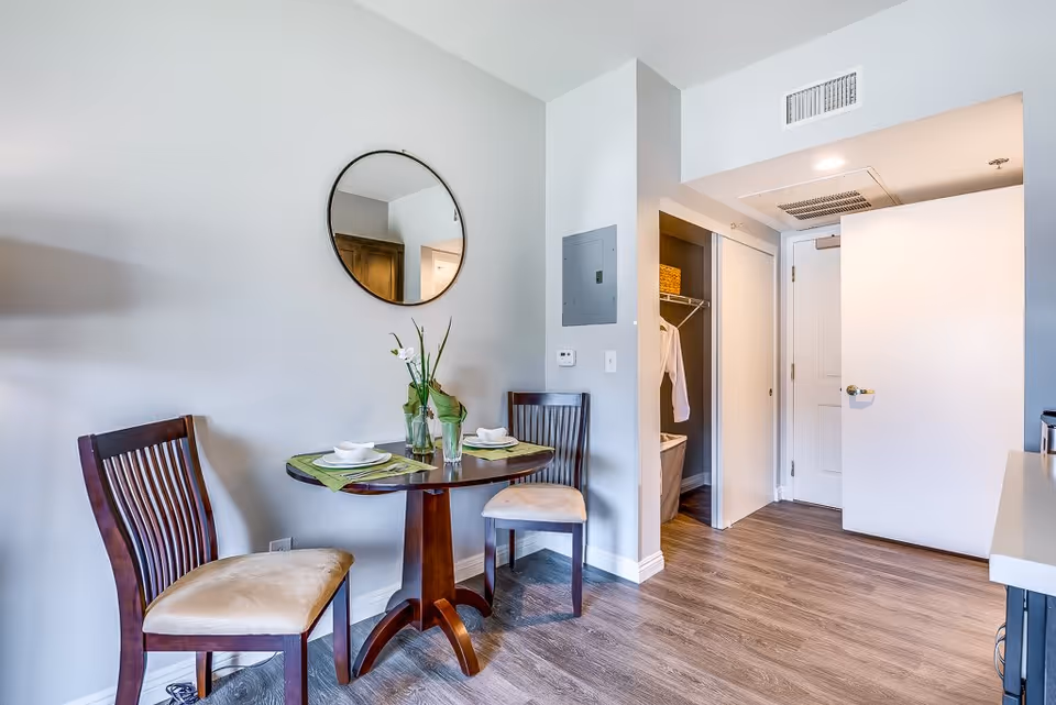 Small dining area with a round wooden table set for two with plates, bowls, and glasses. Two wooden chairs with beige cushions are placed around the table. A round mirror hangs on the light gray wall above the table. To the right, there is an open closet with clothes hanging and a white door leading to another room. The floor is covered with wood-style laminate flooring.