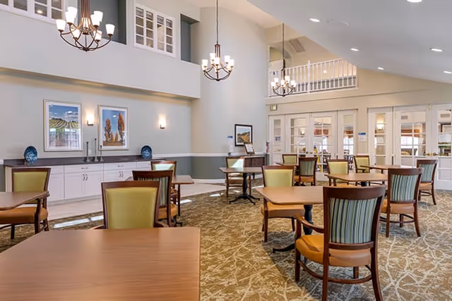 A spacious dining room with multiple wooden tables and chairs arranged neatly on a patterned carpet. The room features high ceilings with hanging chandeliers, light-colored walls with framed artwork, and large windows with white trim. There is a sideboard with decorative items along one wall and a set of double doors leading to another area.