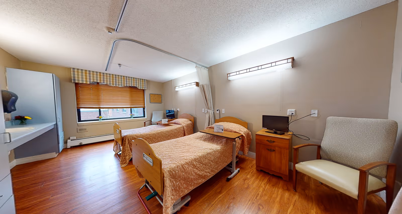 A bright and clean room with two hospital-style beds covered in matching beige bedspreads. Each bed has a small overbed table. There is a wooden nightstand with a small television on it between the beds. A beige armchair is positioned to the right of the nightstand. The room has wooden flooring, a large window with wooden blinds and a plaid valance, and a sink area on the left side. The walls are light-colored, and there are overhead lights above each bed.