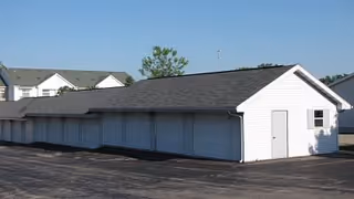 A long white building with a dark shingled roof, multiple garage doors, and a single white door on the side, situated on a paved surface with houses and trees in the background under a clear blue sky.