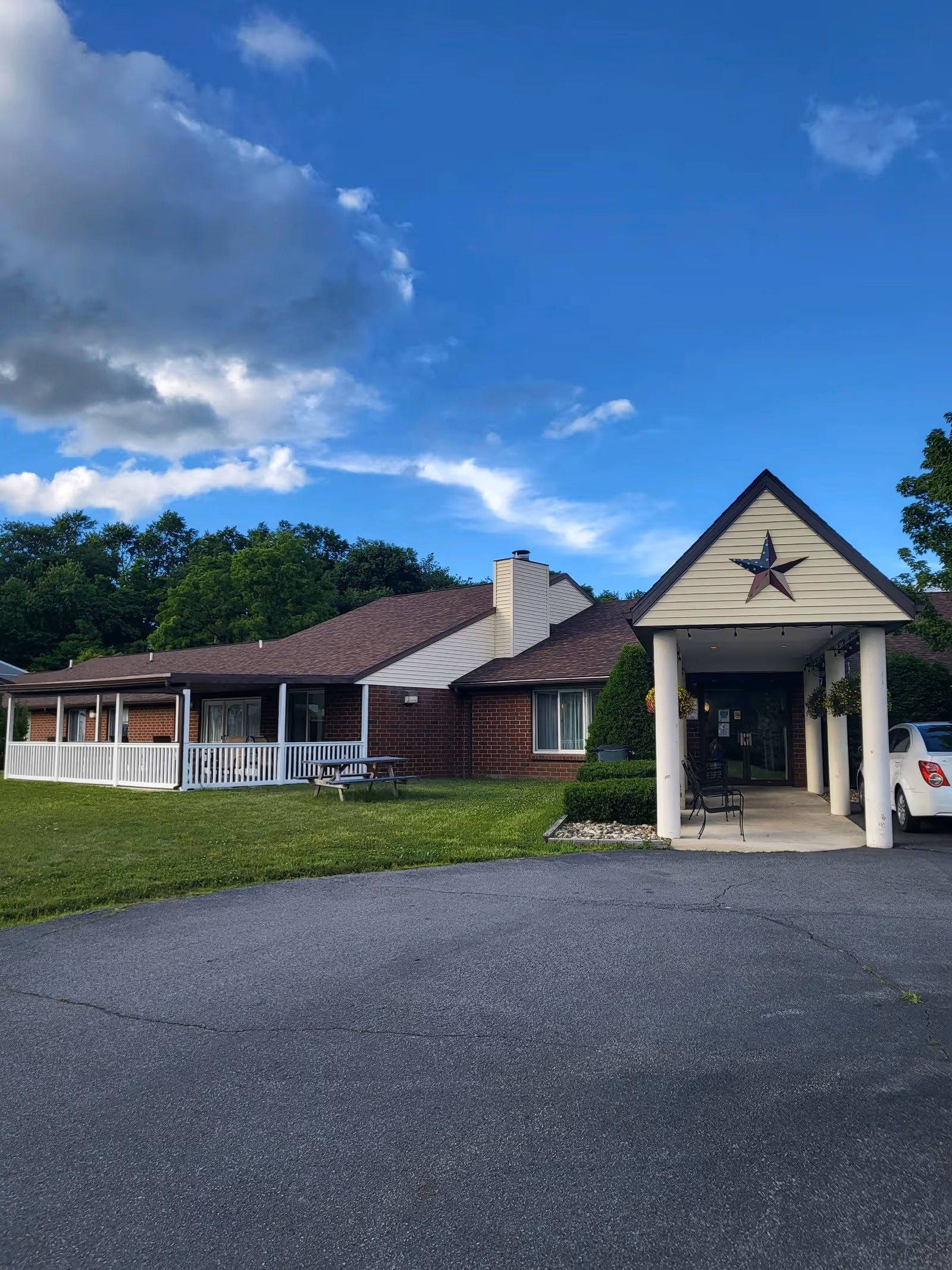 Exterior view of a single-story brick building with a covered entrance supported by white columns. The building has a brown roof and a large star decoration above the entrance. There is a grassy lawn with a picnic table and trees in the background under a partly cloudy blue sky.