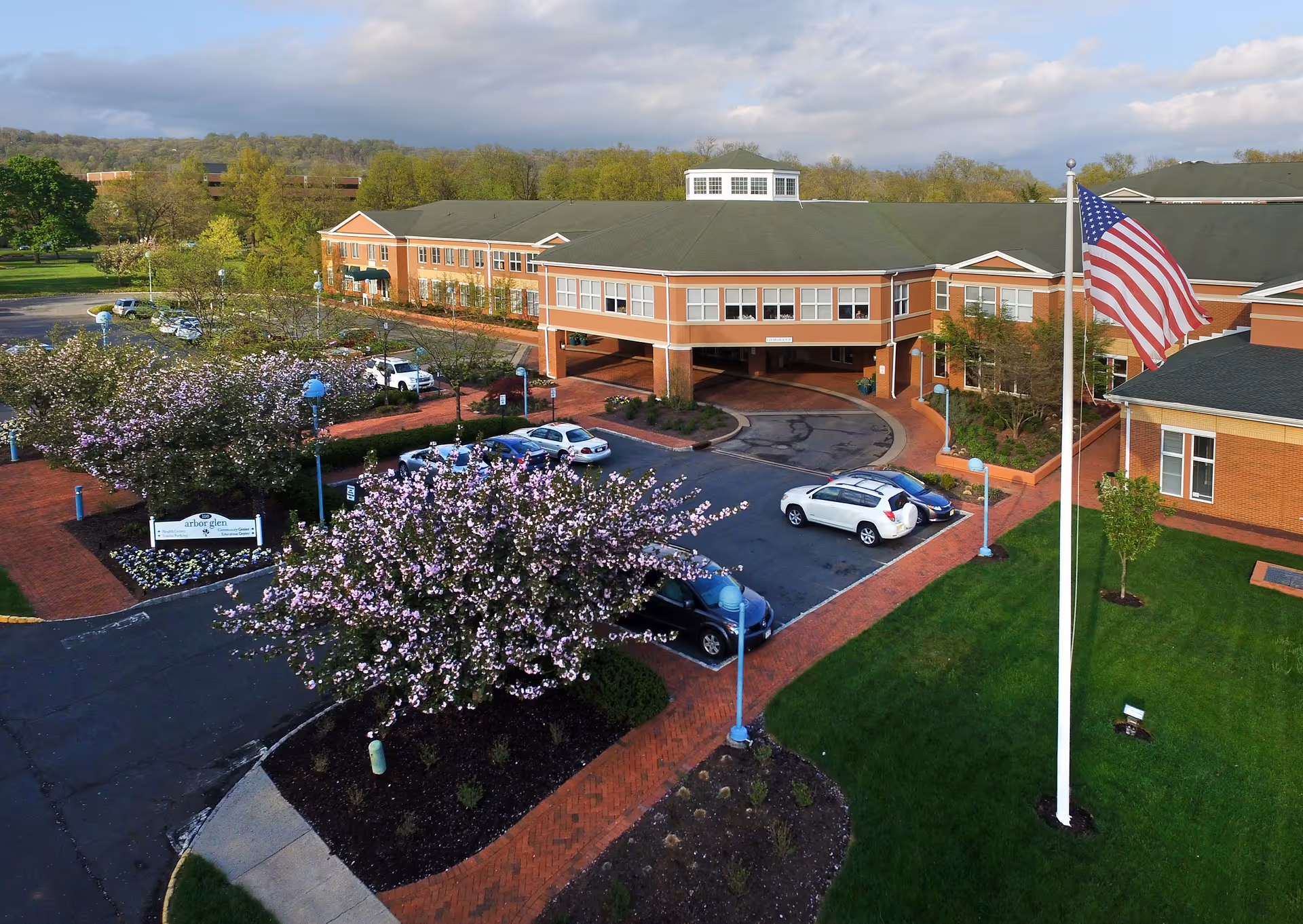 Aerial view of a senior living facility with a large brick building, a circular driveway with parked cars, blooming trees, a well-maintained lawn, and an American flag on a flagpole.