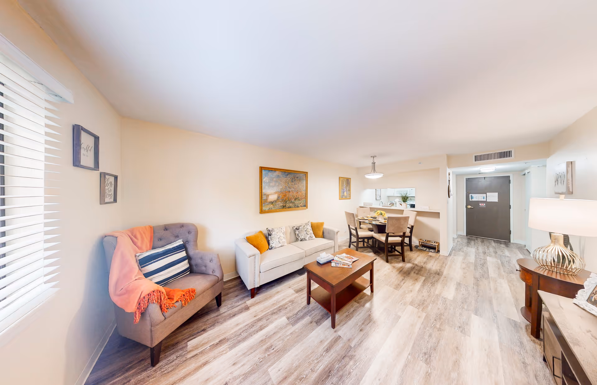 Bright furnished living room and dining area with a sofa, armchair, coffee table, and small dining table in a senior living apartment.