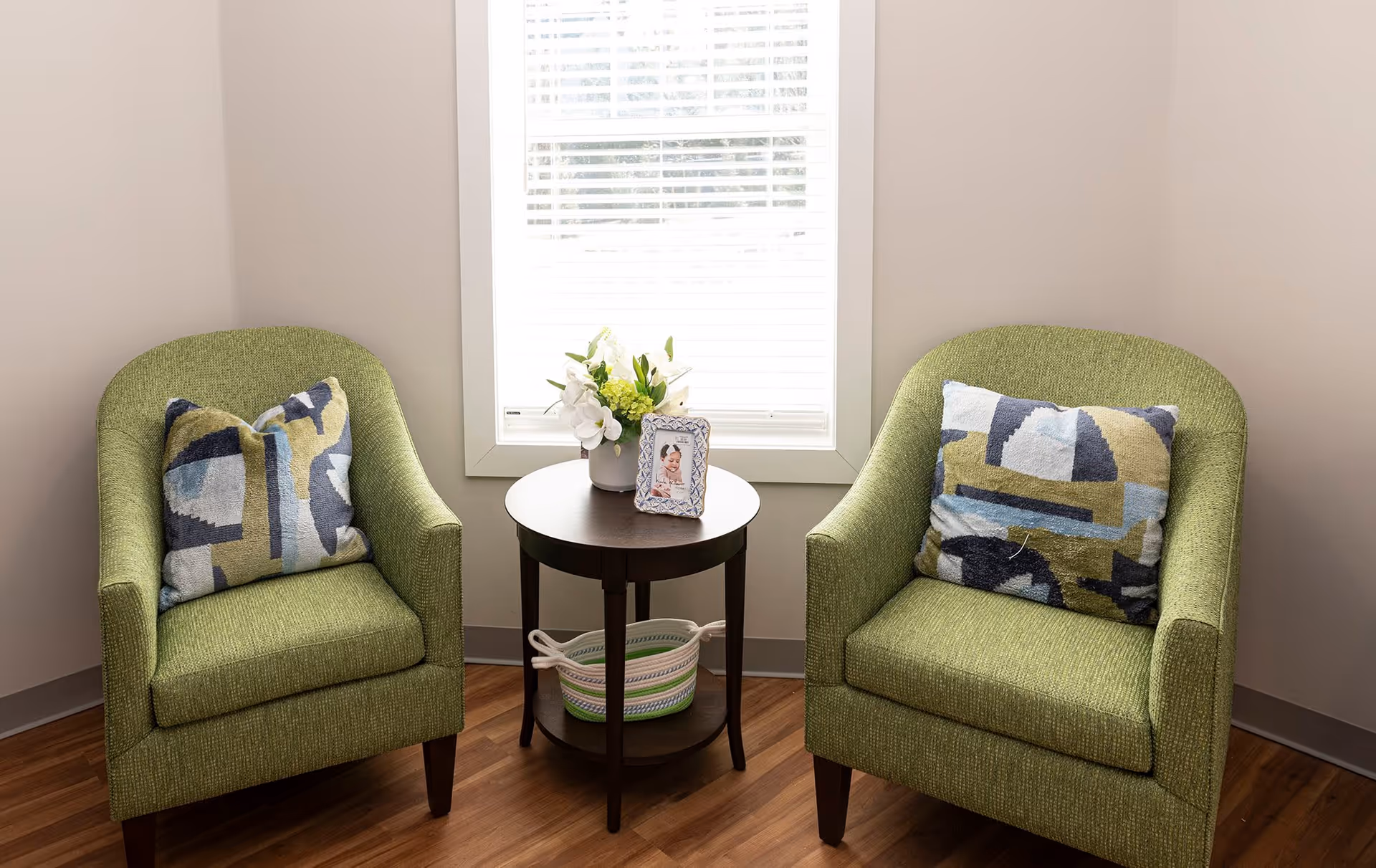 Two green upholstered armchairs with patterned cushions are positioned on either side of a small round wooden table. The table holds a small flower arrangement and a framed photo. Behind the table is a window with white blinds, and the floor is wooden.