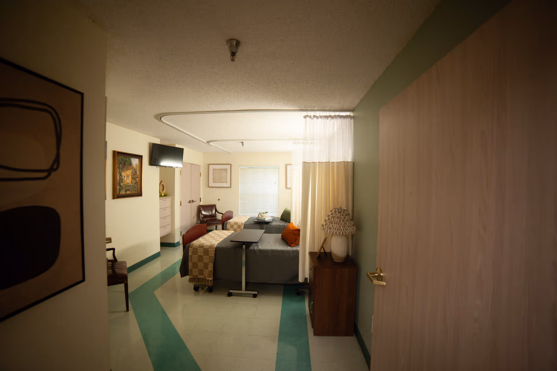 Interior view of a shared resident bedroom with two beds, a privacy curtain, wall-mounted TV, and bedside furniture.
