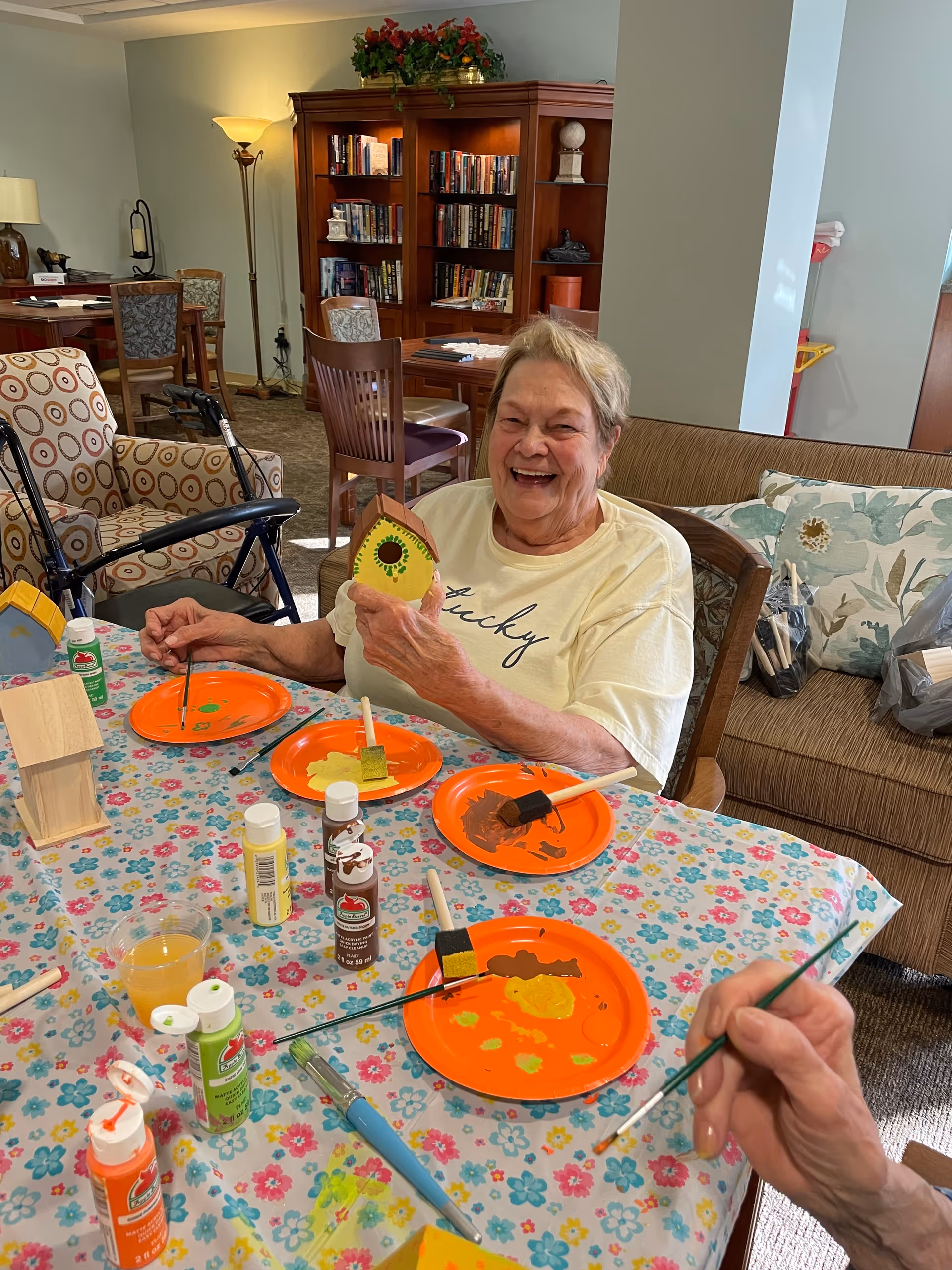 An elderly woman sitting at a table covered with a floral tablecloth, smiling and holding a small painted wooden birdhouse. The table has several orange plates with paint and brushes, along with bottles of paint. In the background, there is a bookshelf filled with books, chairs, and a couch with floral cushions in a cozy room.