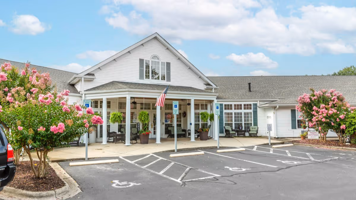 Front exterior view of a single-story building with white siding and a gray roof, featuring a covered entrance with white columns and hanging plants. There are pink flowering bushes on either side of the entrance and several handicap parking spaces in front. The sky is partly cloudy.