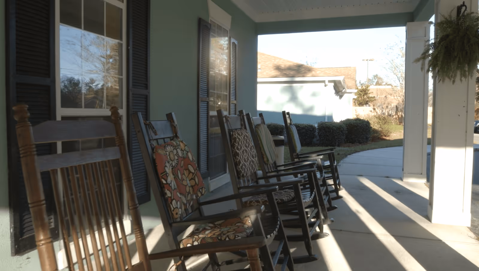 A row of wooden rocking chairs with patterned cushions lined up on a covered porch outside a building, with windows and black shutters visible on the green exterior wall. Sunlight casts shadows on the concrete floor, and there are bushes and a walkway in the background.