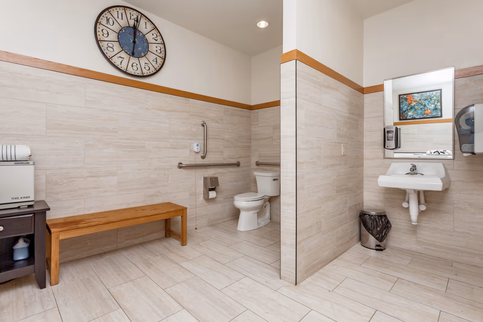 A clean and spacious bathroom with beige tiled walls and floor. The room features a white toilet with grab bars on the walls for accessibility, a white sink with a mirror above it, a soap dispenser, a small trash can, a wooden bench, and a towel warmer on a small dark wooden table. A large clock is mounted on the wall above the bench.