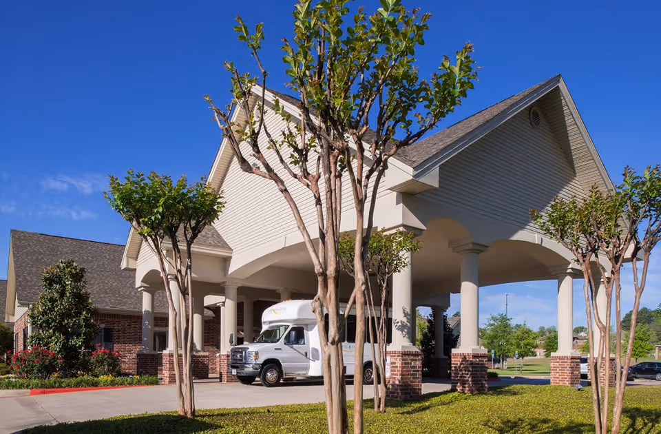 Exterior view of The Heights of Tyler senior living facility entrance with a covered driveway supported by columns, a white shuttle bus parked under the canopy, and landscaped greenery including small trees and bushes under a clear blue sky.