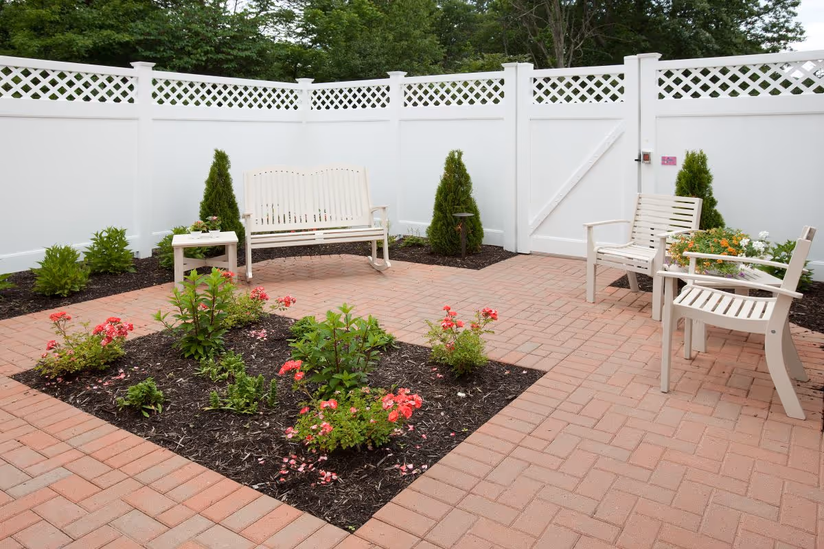 A small outdoor patio area enclosed by a white fence with lattice top. The patio has a brick floor and features a garden bed with blooming pink flowers in the center. There are white wooden benches and chairs arranged around the patio, along with some small green shrubs and potted plants near the fence.