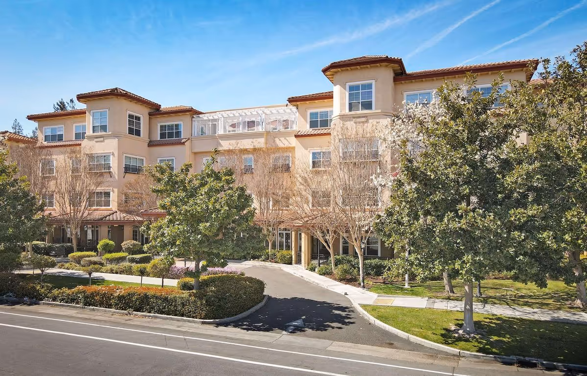 Exterior view of a multi-story senior living facility building with beige walls and red-tiled roof, surrounded by trees and landscaped greenery under a clear blue sky.