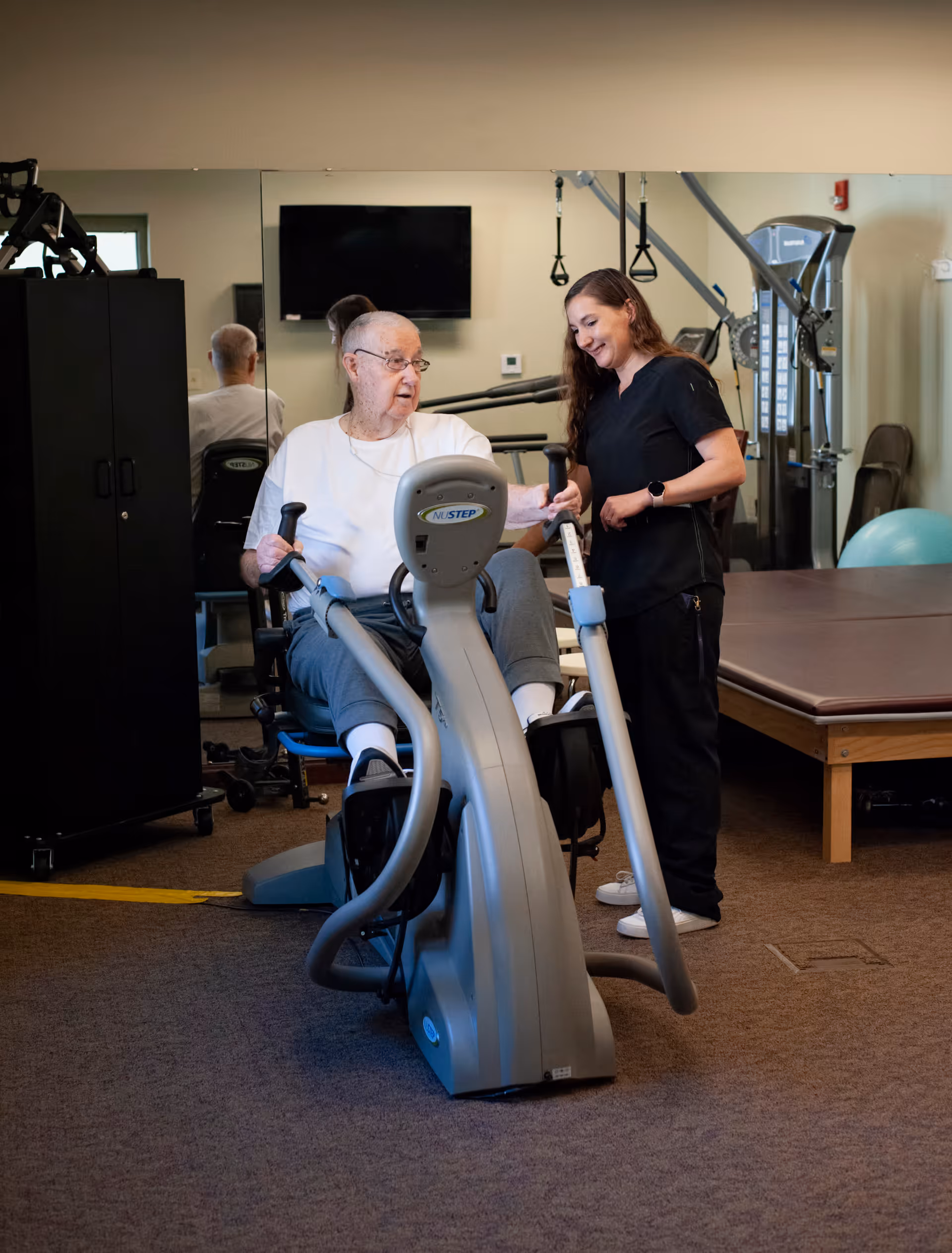 An elderly man using a NuStep exercise machine in a fitness or therapy room, assisted by a female caregiver or therapist who is smiling and providing support. The room has exercise equipment, a large mirror, and a television mounted on the wall.