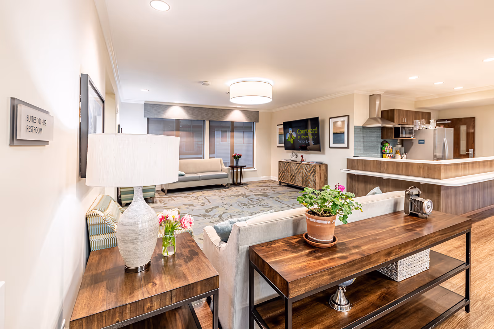 A bright and spacious senior living common area with a beige sofa, wooden tables with decorative items including a lamp, flowers, and a potted plant. The room features a large flat-screen TV mounted on the wall displaying the Courtyard at Mount Tabor Senior Living logo. In the background, there is a modern kitchen area with stainless steel appliances and wooden cabinetry. A sign on the wall indicates directions to suites 100-122 and a restroom.
