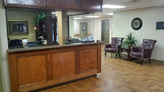Reception area with a wooden front desk and a glass partition. Behind the desk are office supplies and a computer. To the right, there are two maroon leather chairs with white piping, a small wooden table with a plant, and a wall clock above them. The floor is wooden parquet, and the walls are light-colored.