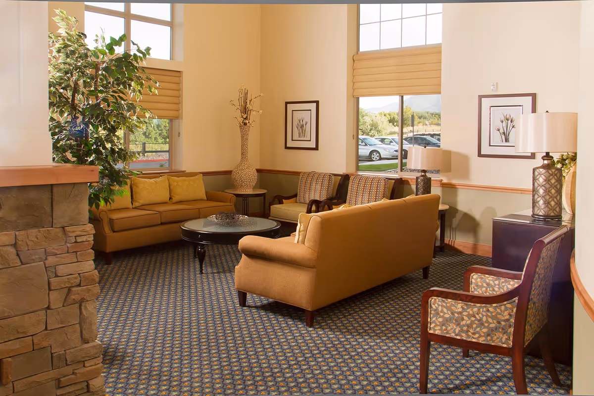 A cozy living room area with two mustard-colored sofas, two patterned armchairs, a round glass coffee table, a large potted plant, two table lamps, framed floral artwork on the walls, and large windows with beige blinds letting in natural light. The room has a patterned carpet and a stone fireplace partially visible on the left.