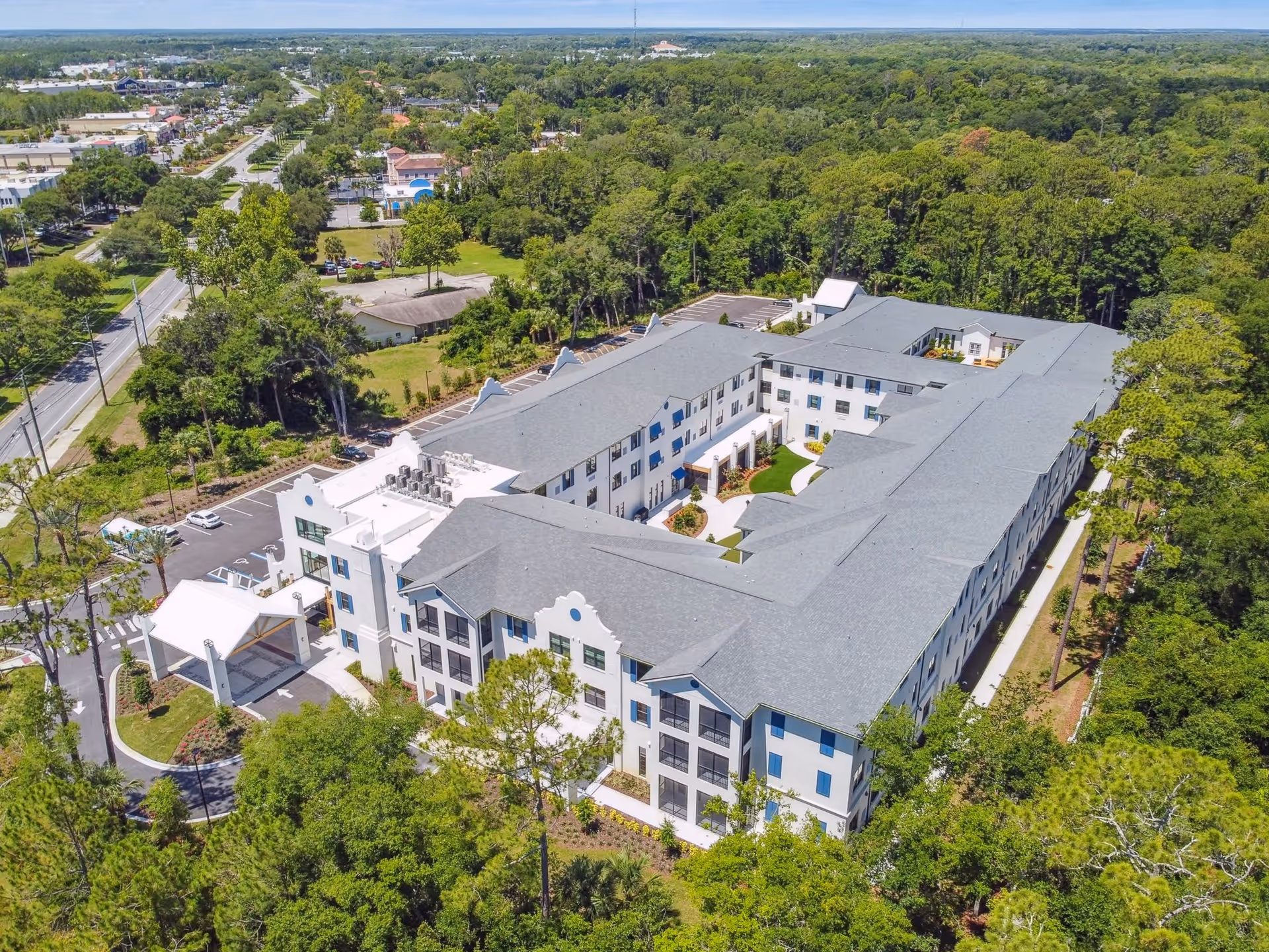 Aerial view of a large, modern senior living facility named Aden Senior Living surrounded by trees and greenery. The building is three stories tall with a gray roof and white exterior walls. There is a covered entrance at the front and a courtyard in the center of the building. The facility is located near a road with parking spaces visible around the building.