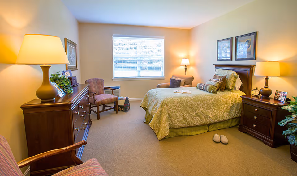 A cozy bedroom with a large window letting in natural light. The room features a bed with green patterned bedding, two bedside tables with lamps, a dresser with a lamp and decorative items, two chairs, and a small plant in the corner. The walls are painted a soft yellow, and there are framed pictures above the bed.
