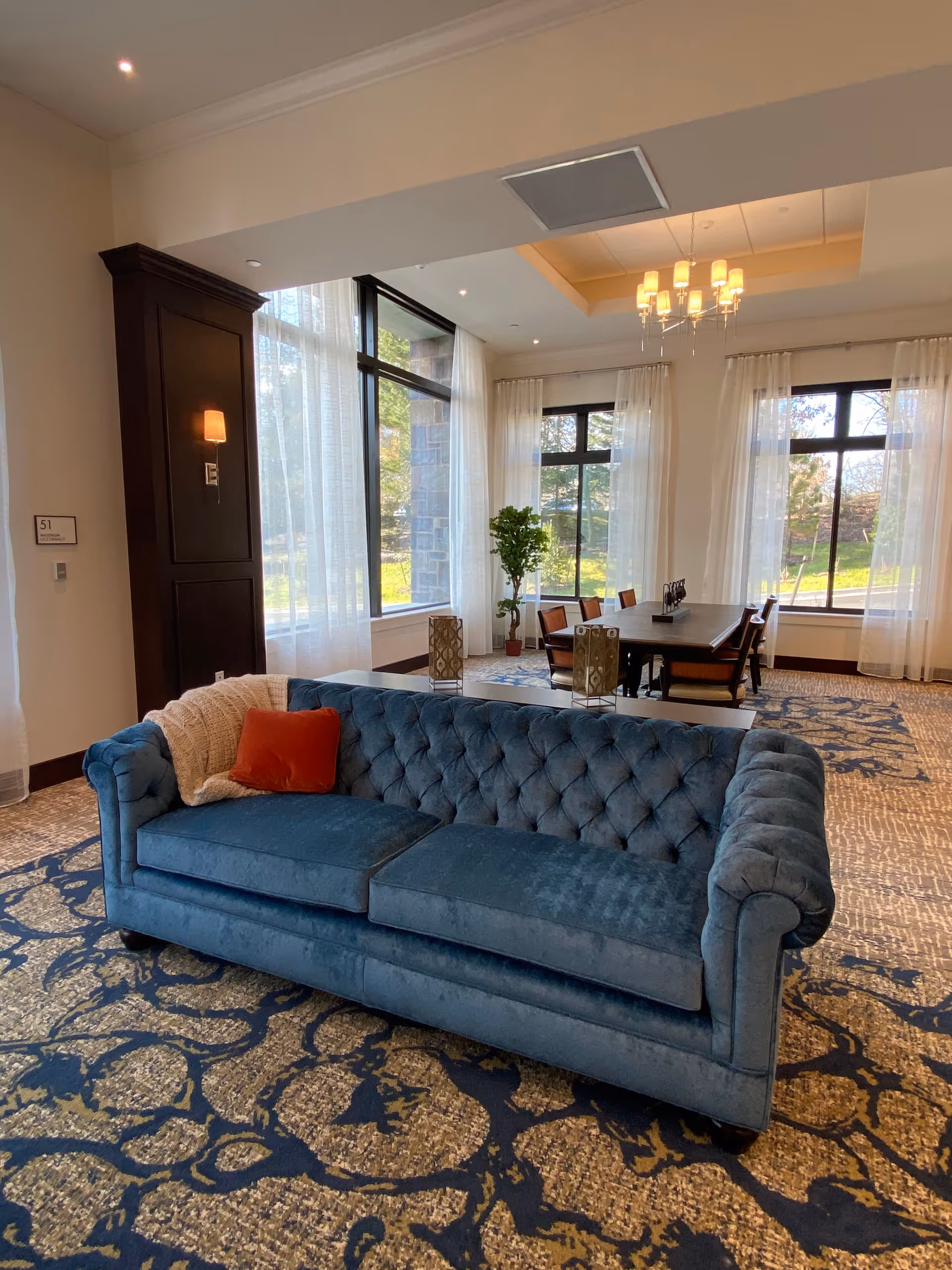A tufted blue velvet sofa sits on a patterned carpet in a bright communal lounge with large windows, sheer curtains, and a dining table in the background.