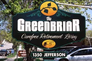A large green and yellow sign for The Greenbriar Carefree Retirement Living located at 1350 Jefferson, with trees and parked cars visible in the background.