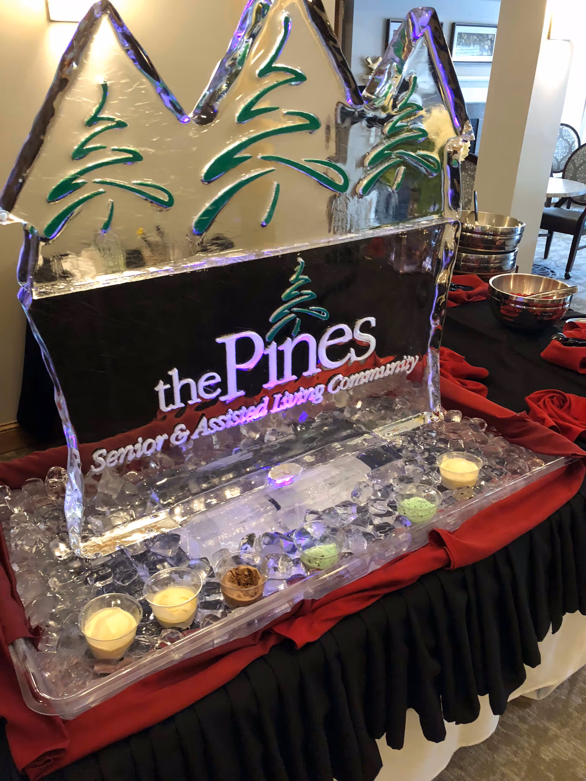 An ice sculpture with green pine tree designs and the text 'the Pines Senior & Assisted Living Community' carved into it, displayed on a table covered with a black and red cloth. The table also holds small cups of various colored ice cream or dessert samples and metal bowls in the background.
