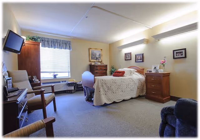 A cozy senior living bedroom with a single bed covered in a white quilt, wooden furniture including a dresser, nightstand, and chairs, a wall-mounted TV, and a window with blinds and a valance letting in natural light.
