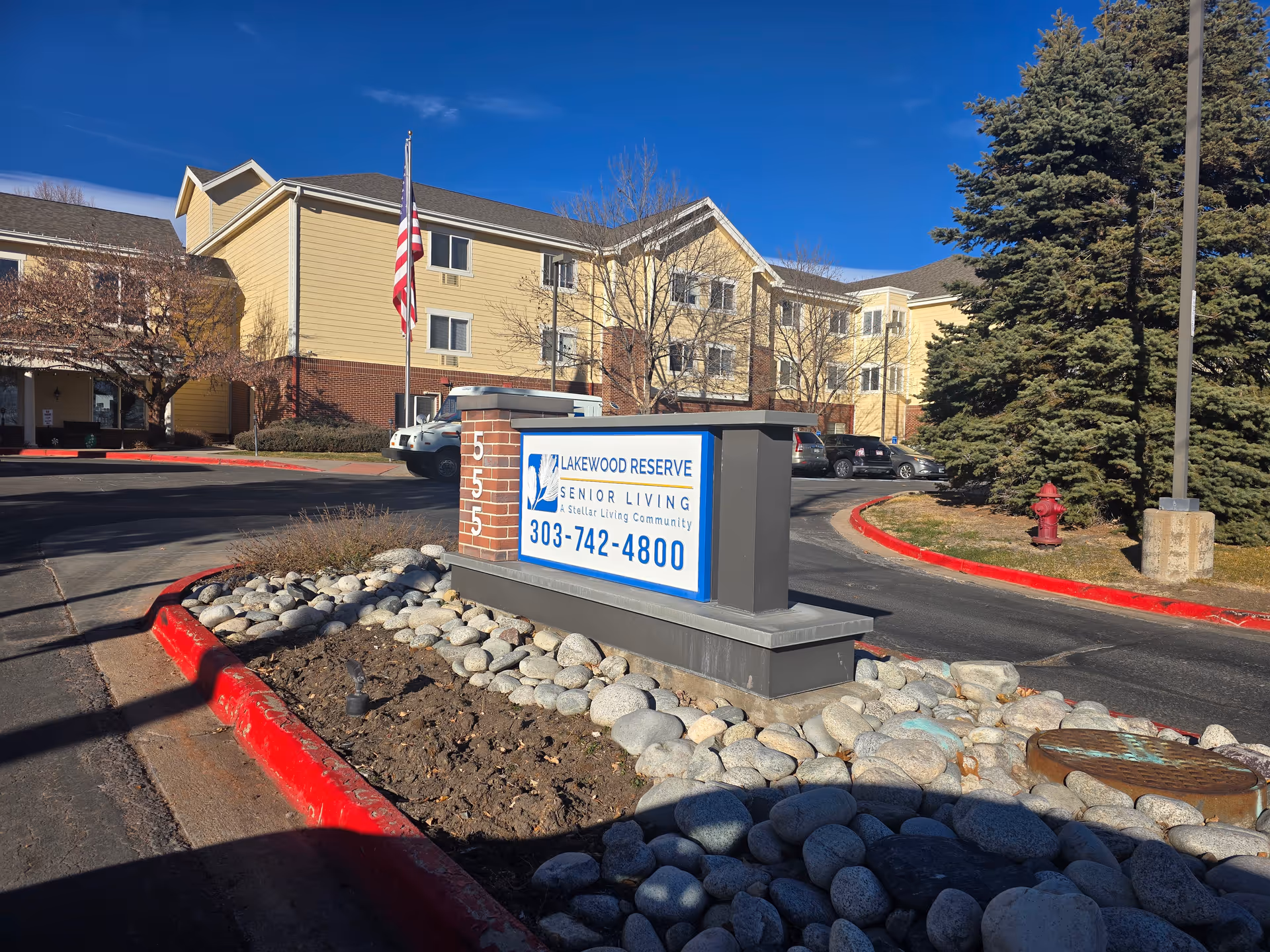 Exterior view of Lakewood Reserve Assisted Living & Memory Care facility with a large sign displaying the facility name, phone number, and address number 5555. The building is three stories tall with yellow siding and brick accents. There is an American flag on a pole near the sign, several parked cars, trees, and a clear blue sky.
