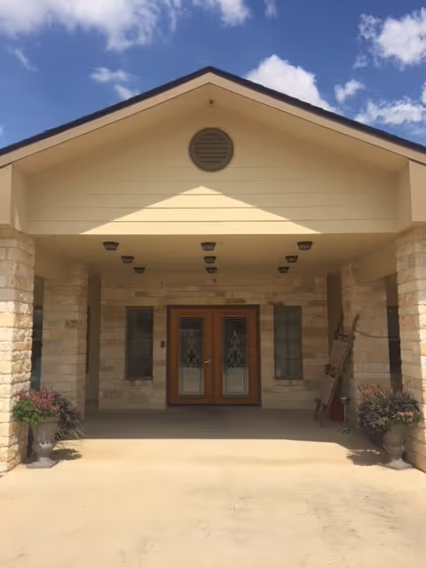 Entrance of a building with a covered porch supported by stone pillars, double wooden doors with decorative glass panels, two windows on either side, potted plants near the pillars, and a clear blue sky with some clouds above.
