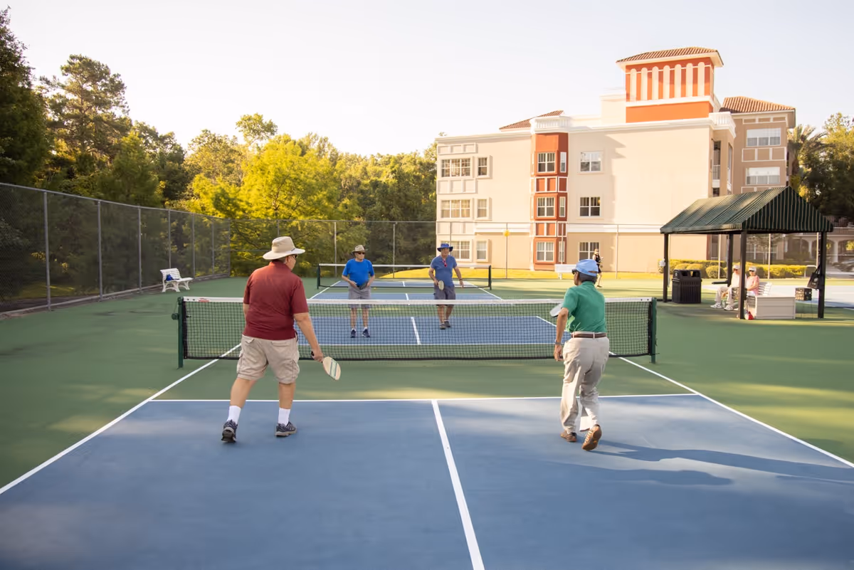 Four elderly men playing pickleball on an outdoor court at a senior living facility, with a multi-story building and trees in the background. Two men are on each side of the net, holding paddles and wearing casual clothing and hats. There is a covered seating area and benches nearby.