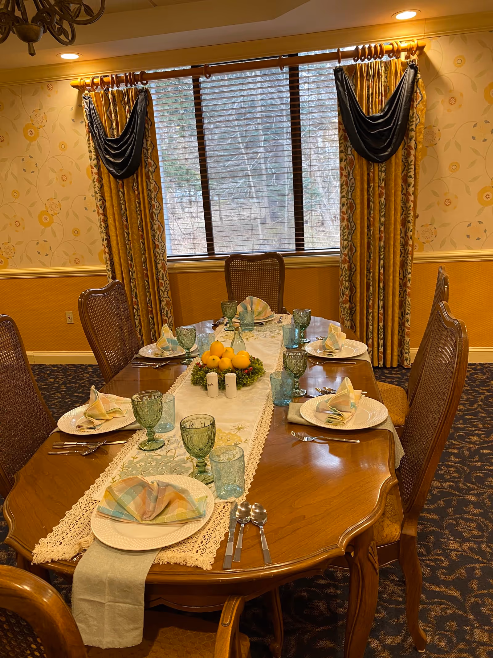 A formally set wooden dining table with plates, folded napkins, green glassware and a fruit centerpiece in a room with patterned curtains and a large window.