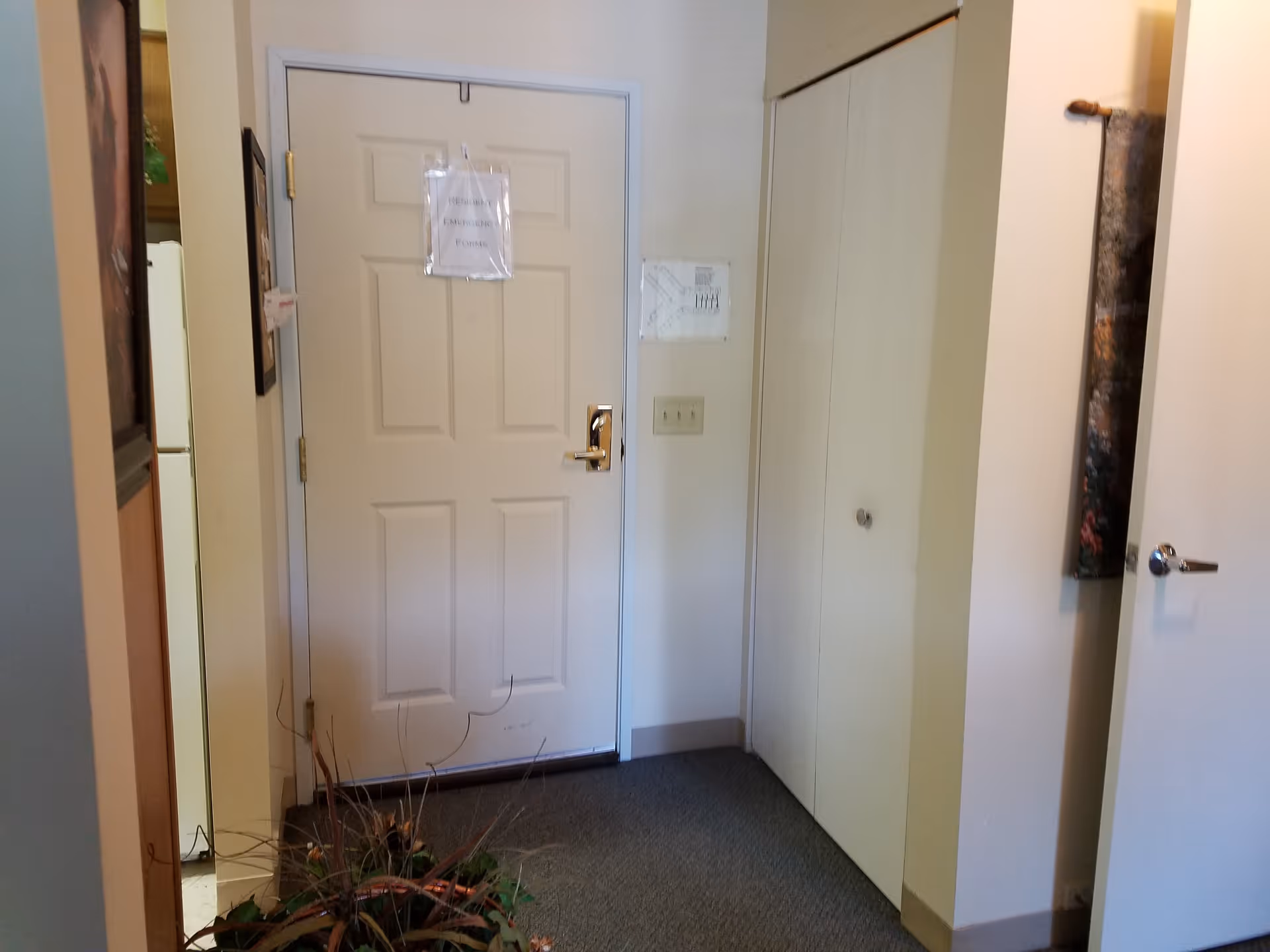 Interior view of a hallway in a senior living facility with a closed white door labeled 'Resident Emergency Forms', a closet with double doors, a partially visible open door on the right, a carpeted floor, and a plant in the foreground.