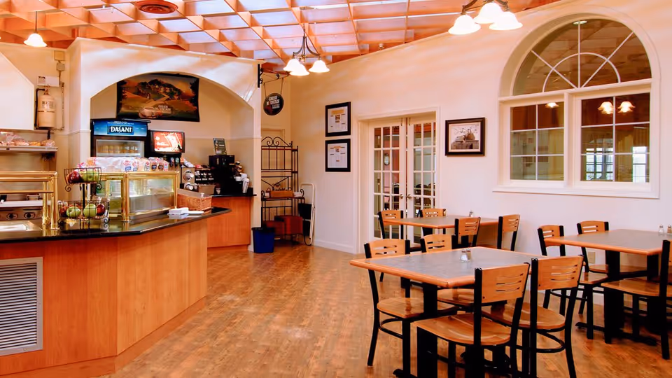 A bright dining area with wooden tables and chairs, a counter with a glass display case containing food items, a coffee machine, and a beverage cooler. The room has a wooden floor, white walls, and a ceiling with a grid pattern and hanging lights. There are framed pictures on the walls and a large arched window.