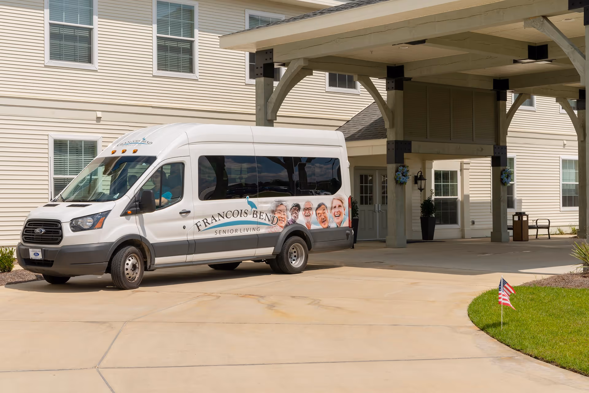 A white shuttle van with 'Francois Bend Senior Living' branding and images of smiling seniors is parked in front of the entrance to a senior living facility building with beige siding and white-framed windows. The entrance has a covered drop-off area supported by wooden beams, and there is a small American flag planted in the grass nearby.