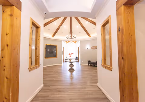 A bright hallway with wooden beams on the ceiling and wooden framed windows on both sides. The floor is light wood, and at the end of the hallway there is a round pedestal table with a vase of flowers. The walls are painted white, and there is a chandelier hanging from the ceiling.