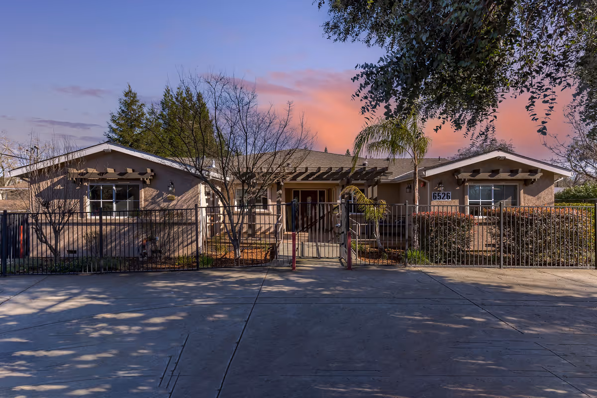 Front exterior view of a single-story senior living facility building with a gated entrance, trees, and bushes in front, under a colorful sunset sky.