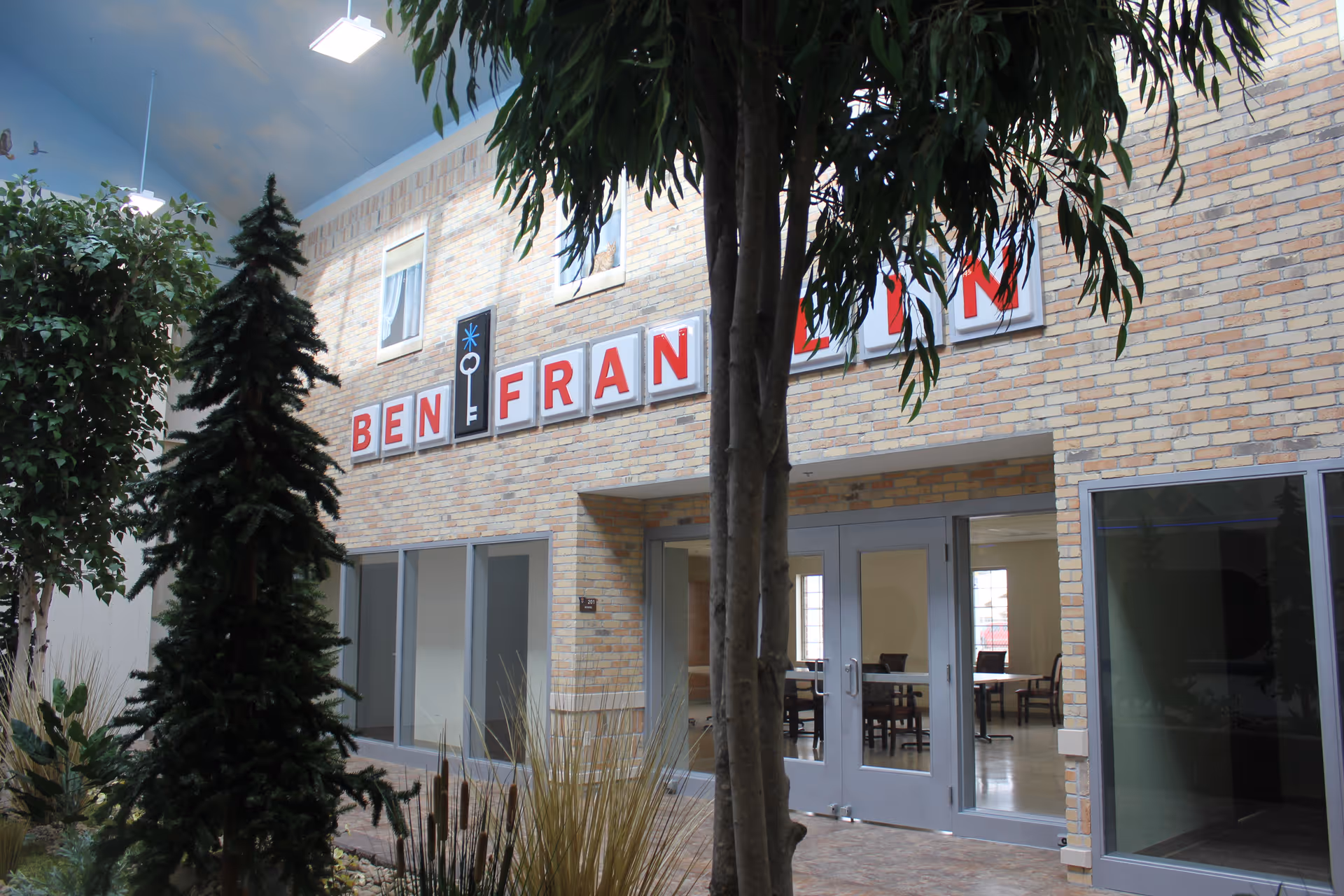 Indoor view of a facility area with a brick wall featuring the sign 'BEN FRANKLIN' above double glass doors. There are several artificial trees and plants in the foreground, and a room with tables and chairs is visible through the glass doors.