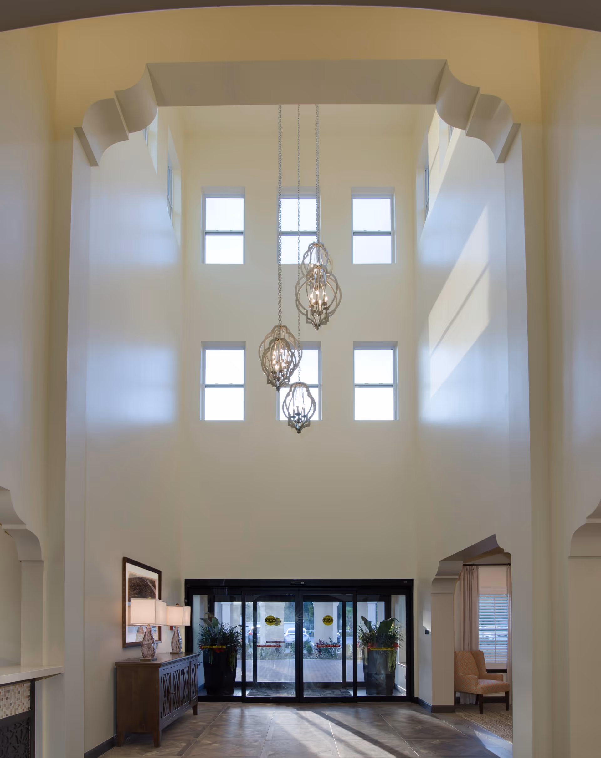 Spacious and well-lit lobby area with high ceilings, multiple windows, and three decorative hanging light fixtures. There is a wooden console table with two lamps and a framed picture on the left side, and large glass automatic doors at the far end. The walls are painted in a light cream color, and there are architectural details around the ceiling and doorways.