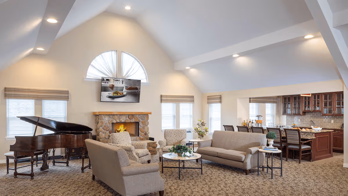 A spacious and well-lit living room area with a high vaulted ceiling, featuring a stone fireplace with a fire burning, a flat-screen TV mounted above it, a grand piano near the windows, two armchairs, a sofa, a loveseat, and a round coffee table with a plant. In the background, there is a kitchen area with a granite countertop island and several bar stools.