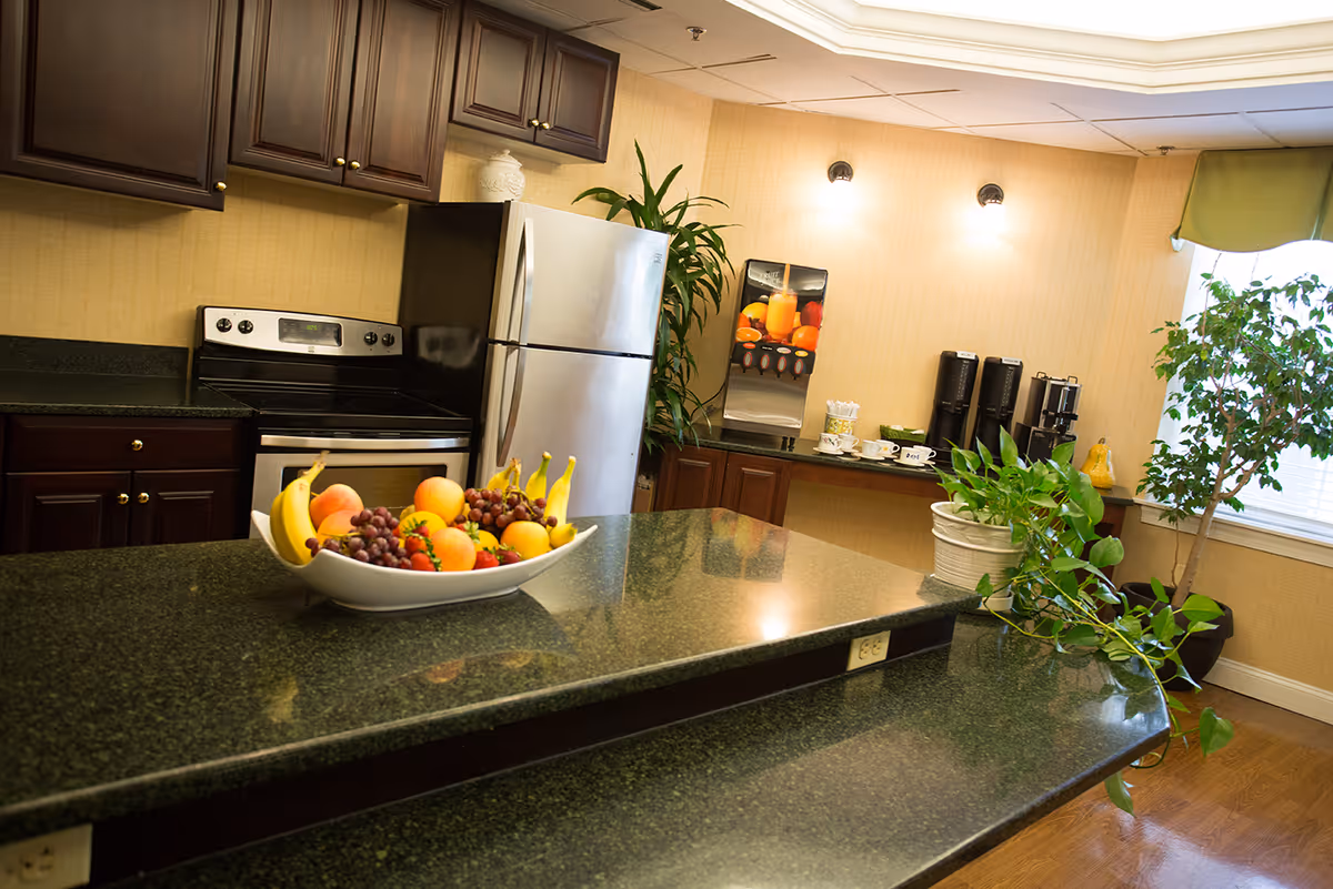 Interior view of a kitchen area with dark wood cabinets, a stainless steel refrigerator and stove, a countertop with a bowl of fresh fruit including bananas, grapes, apples, and strawberries. In the background, there is a beverage dispenser, coffee machines, cups, and green plants near a window with a green valance.