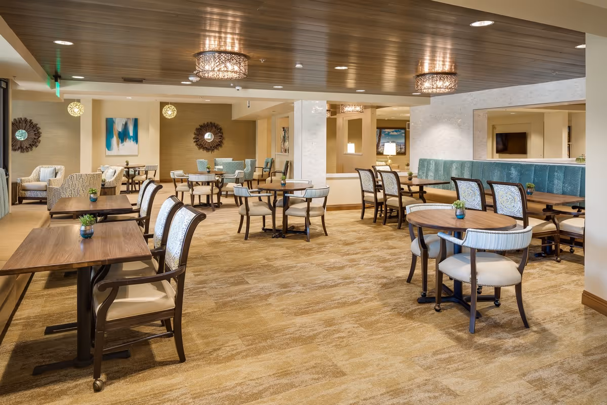 A spacious and well-lit dining area in a senior living facility with multiple wooden tables and cushioned chairs arranged neatly. The room features a beige carpet, decorative wall mirrors, modern light fixtures on the ceiling, and artwork on the walls. There are small potted plants on each table adding a touch of greenery.