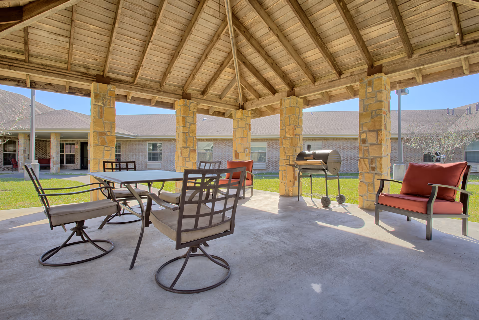 Outdoor covered patio area with a wooden roof supported by stone pillars, featuring a table with four cushioned chairs and two cushioned armchairs. A barbecue grill is also visible under the patio. The background shows a building with brick walls and windows, and a grassy lawn.