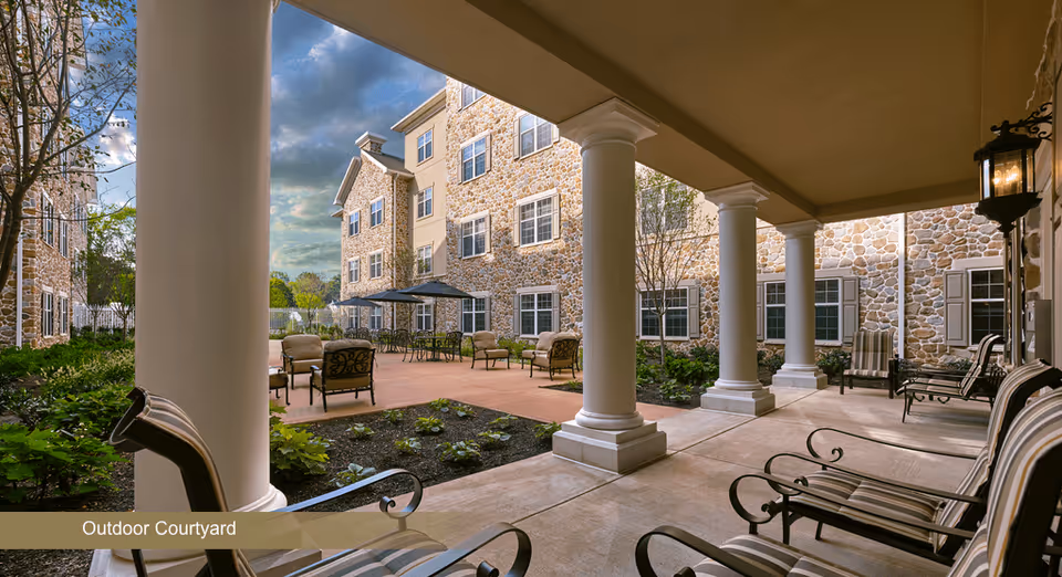 Outdoor courtyard area of a senior living facility with stone building walls, multiple seating areas including cushioned chairs and tables with umbrellas, surrounded by greenery and trees under a partly cloudy sky.