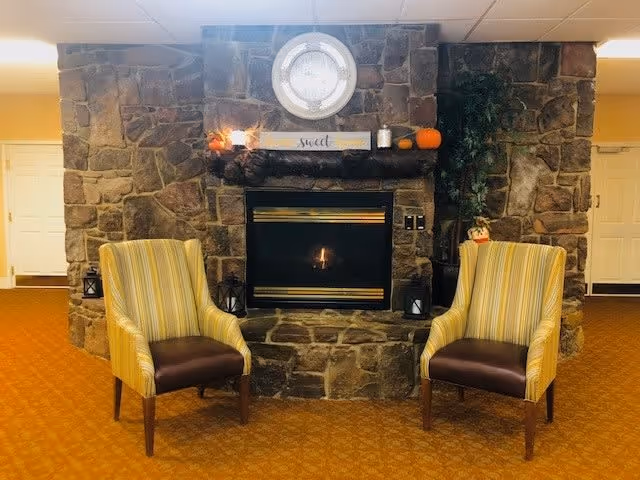 Two striped armchairs with brown seats are placed in front of a stone fireplace with a lit fire. Above the fireplace is a round clock and a wooden sign that says 'Home Sweet Home' along with small decorative pumpkins and candles. The room has a warm, cozy ambiance with orange carpet and yellow walls.