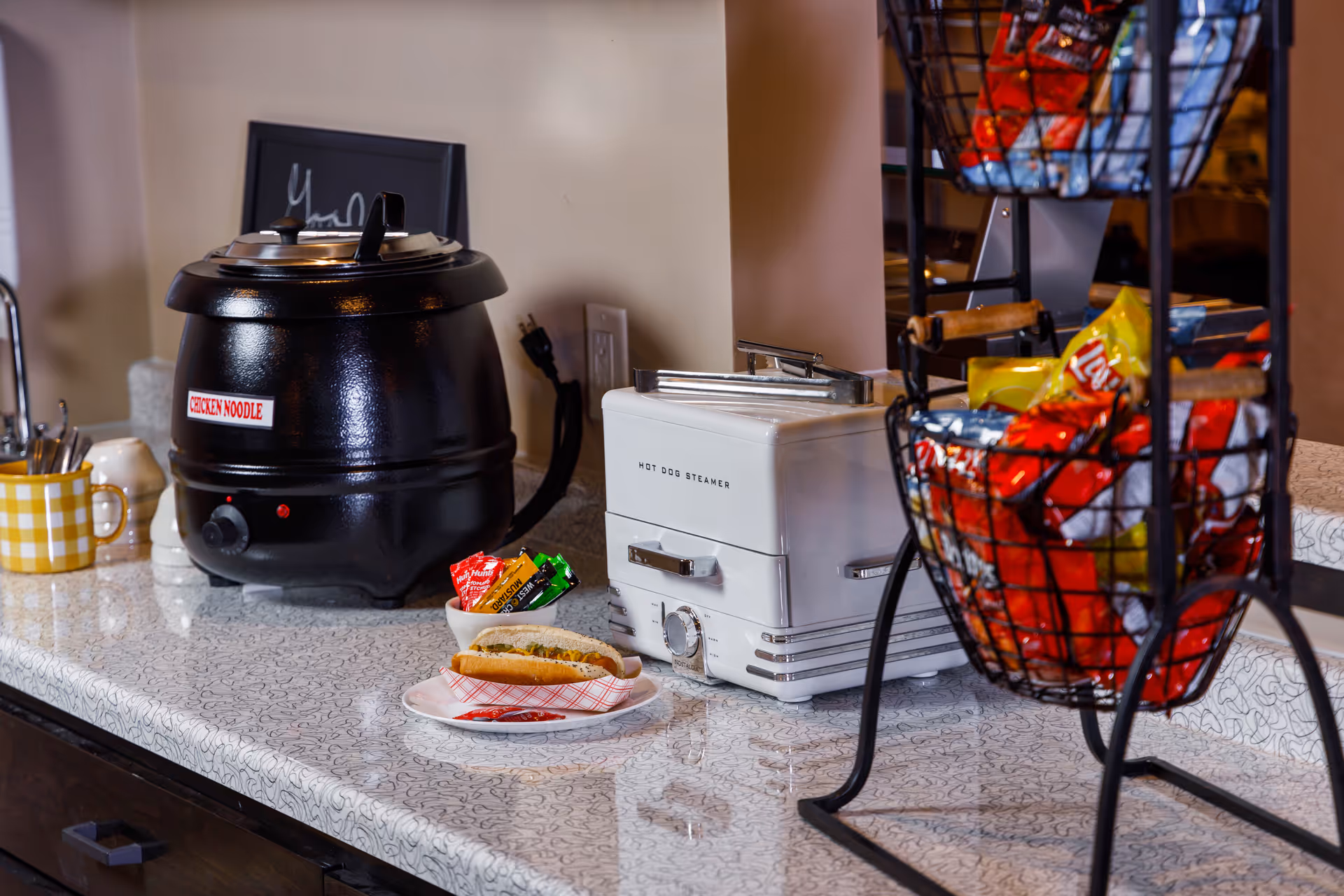 Countertop serving area with a soup kettle labeled "CHICKEN NOODLE", a hot dog steamer, a plate with a hot dog, and baskets of snack chips.