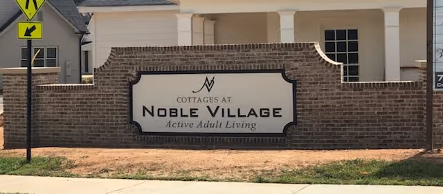 Brick entrance sign that reads "Cottages at Noble Village - Active Adult Living" in front of a residential building.