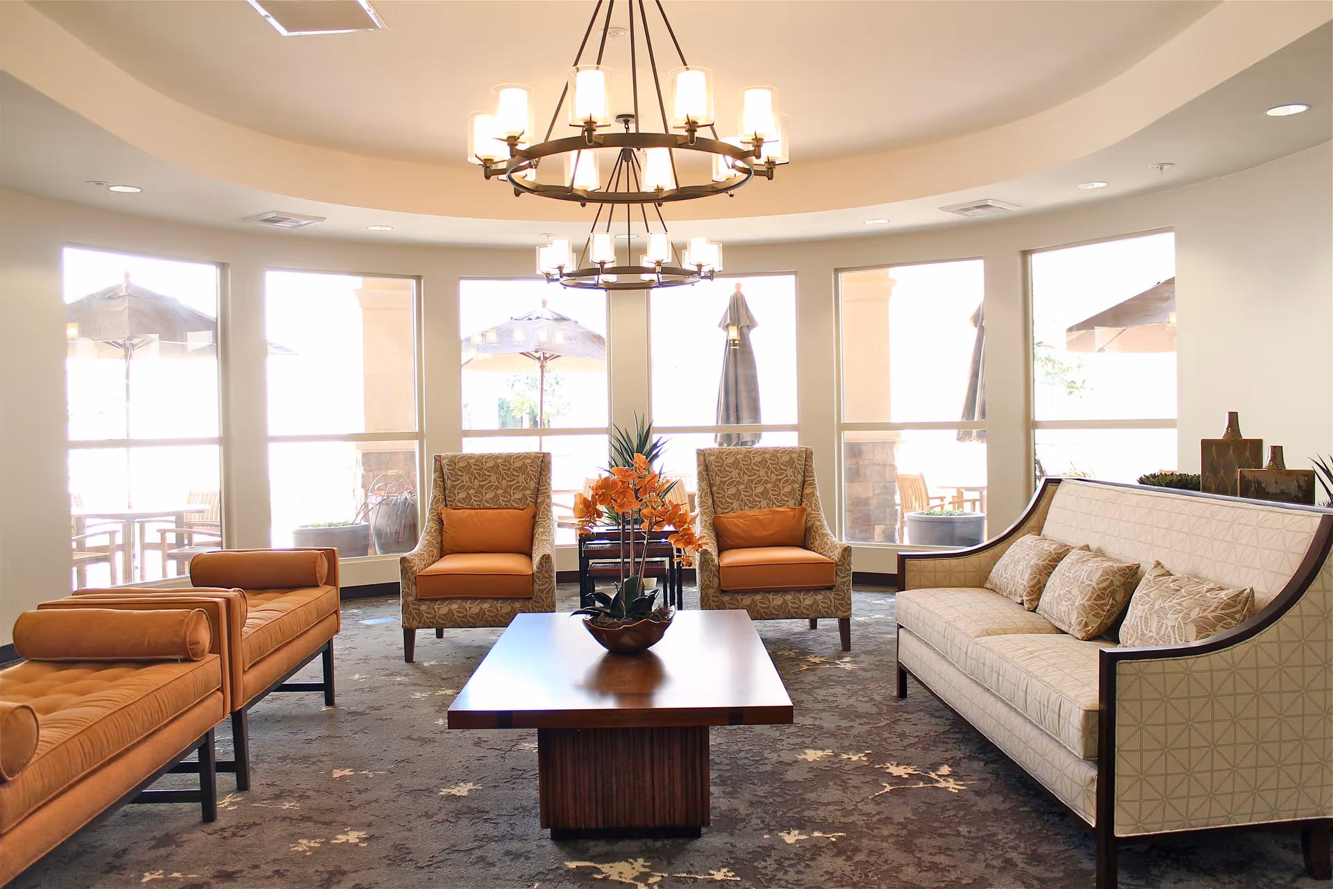 A bright and spacious living room with large windows letting in natural light. The room features two patterned armchairs with orange cushions, a beige sofa with matching pillows, a wooden coffee table with an orange flower arrangement, and a brown cushioned bench. Two circular chandeliers hang from the ceiling, and outside the windows, patio umbrellas and outdoor seating are visible.