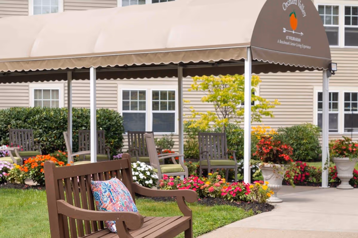 Outdoor seating area at Benchmark at Orchard Valley featuring wooden benches and chairs with cushions, surrounded by colorful flower beds and greenery, under a beige canopy with the Orchard Valley logo and text.
