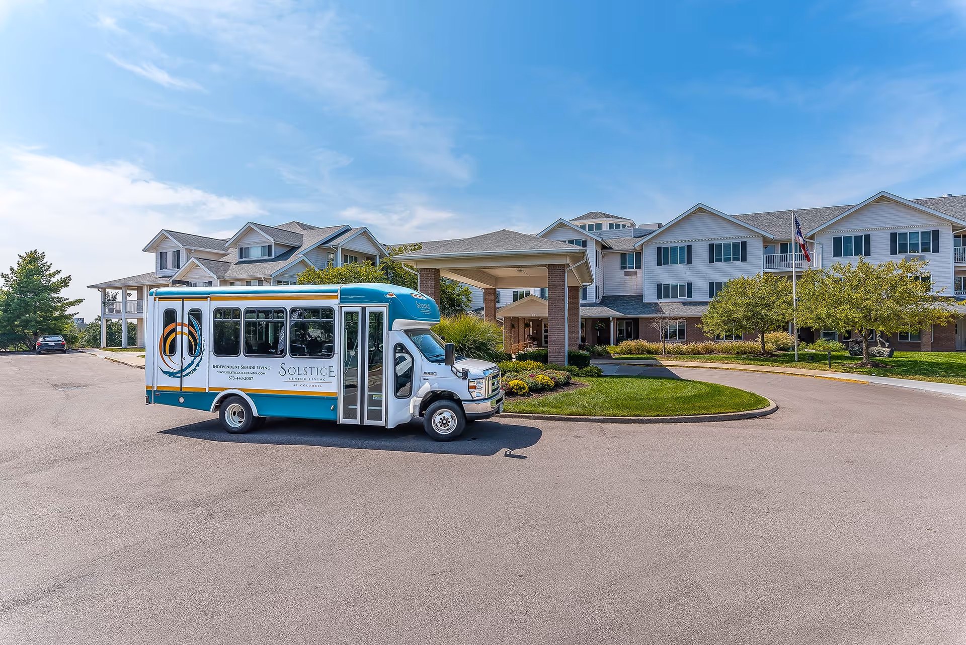 Front entrance of Solstice Senior Living at Columbia with a shuttle bus parked in the driveway in front of a multi-story building under a blue sky.