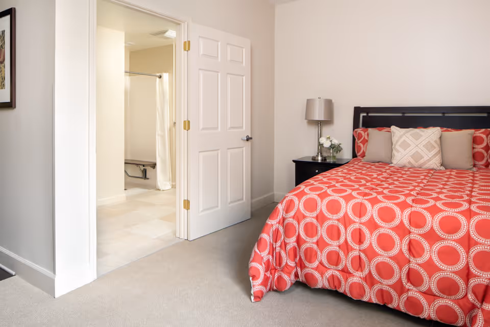A bedroom with a bed covered in a red and white circular patterned bedspread, three pillows, a black nightstand with a lamp and a small flower arrangement. An open door reveals a bathroom with a shower and a white curtain.